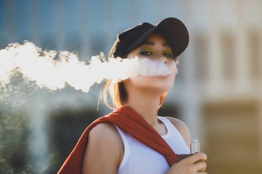 Pretty young hipster asian woman in black hat vape ecig, vaping device at the sunset. Toned image